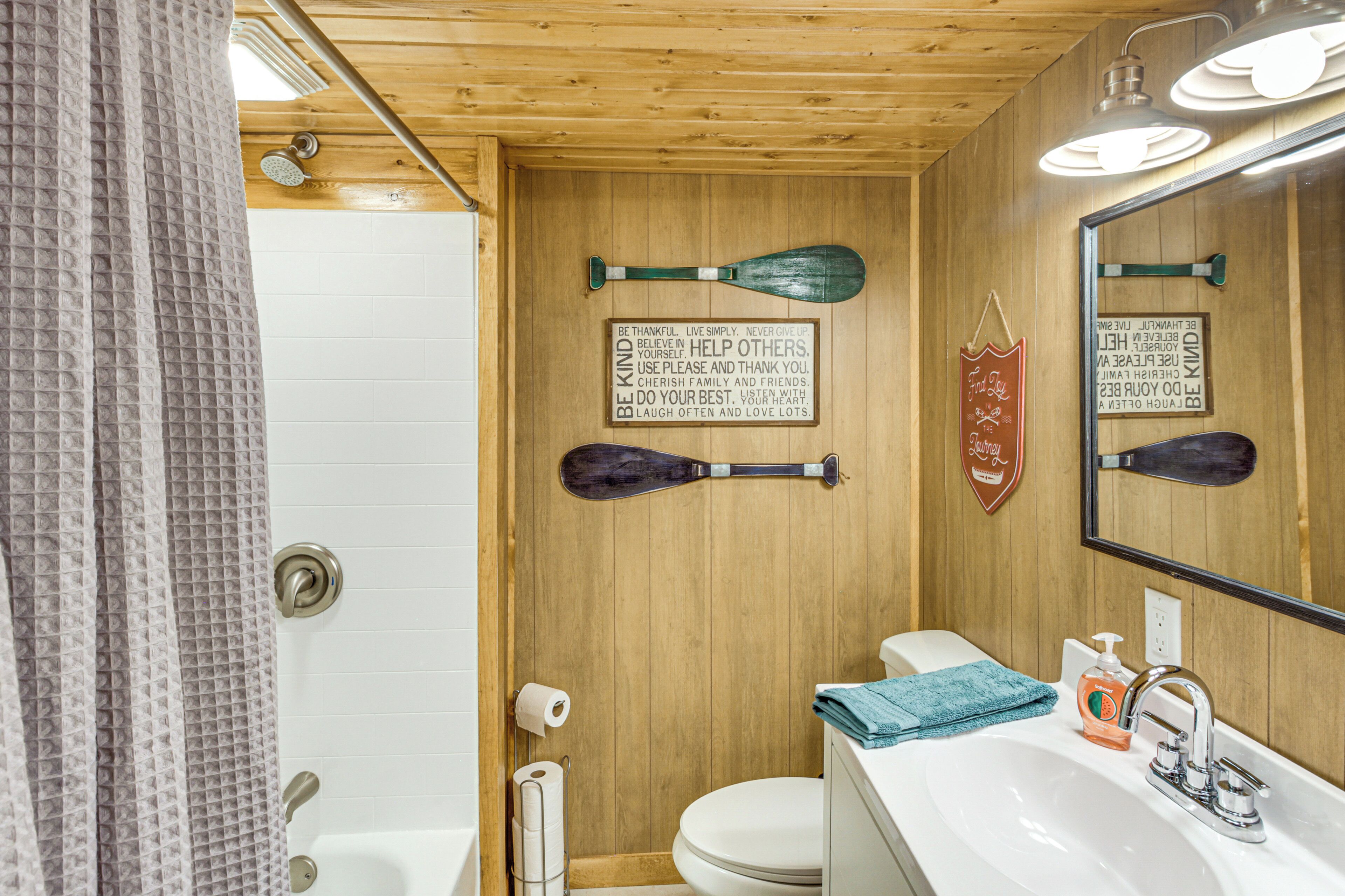Un baño con paredes de madera, una bañera blanca, un inodoro y un lavabo con espejo. Hay dos letreros con forma de remo en la pared y una toalla sobre el lavabo.