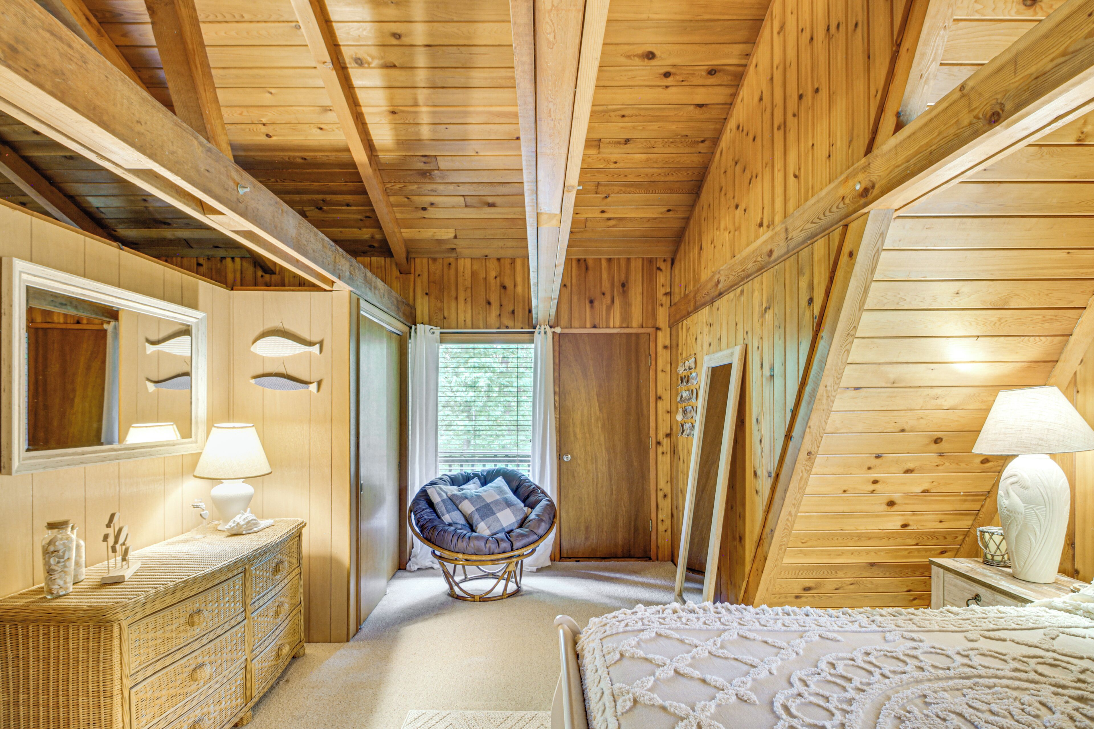 A wooden cabin interior with a bed, dresser, chair, and a window providing natural light.
