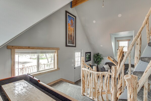 A spacious loft with a high ceiling, a large window, a wooden staircase, and a cozy seating area.