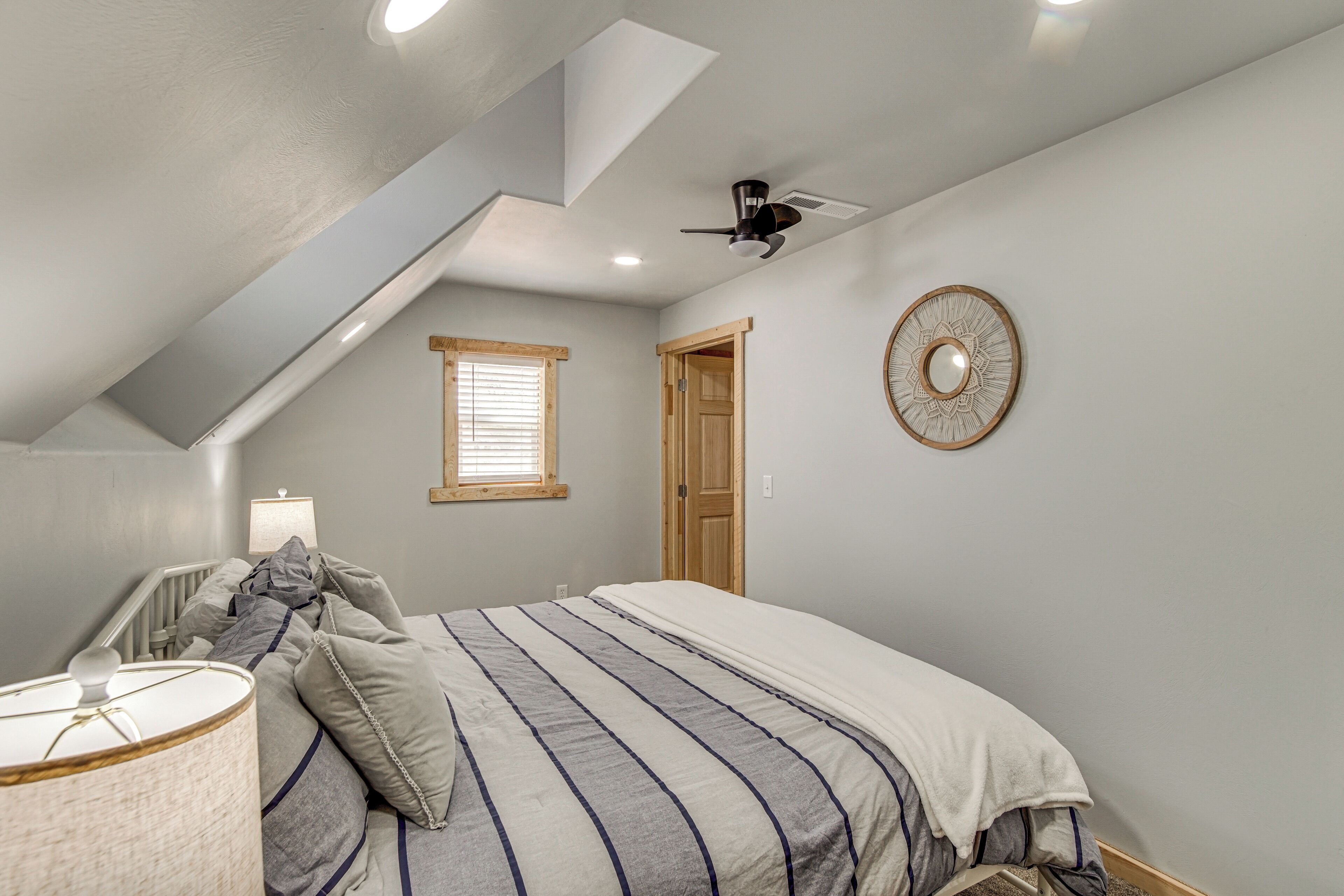 A bedroom with a sloped ceiling, a bed with striped bedding, a ceiling fan, a window with blinds, and a decorative wall plate.