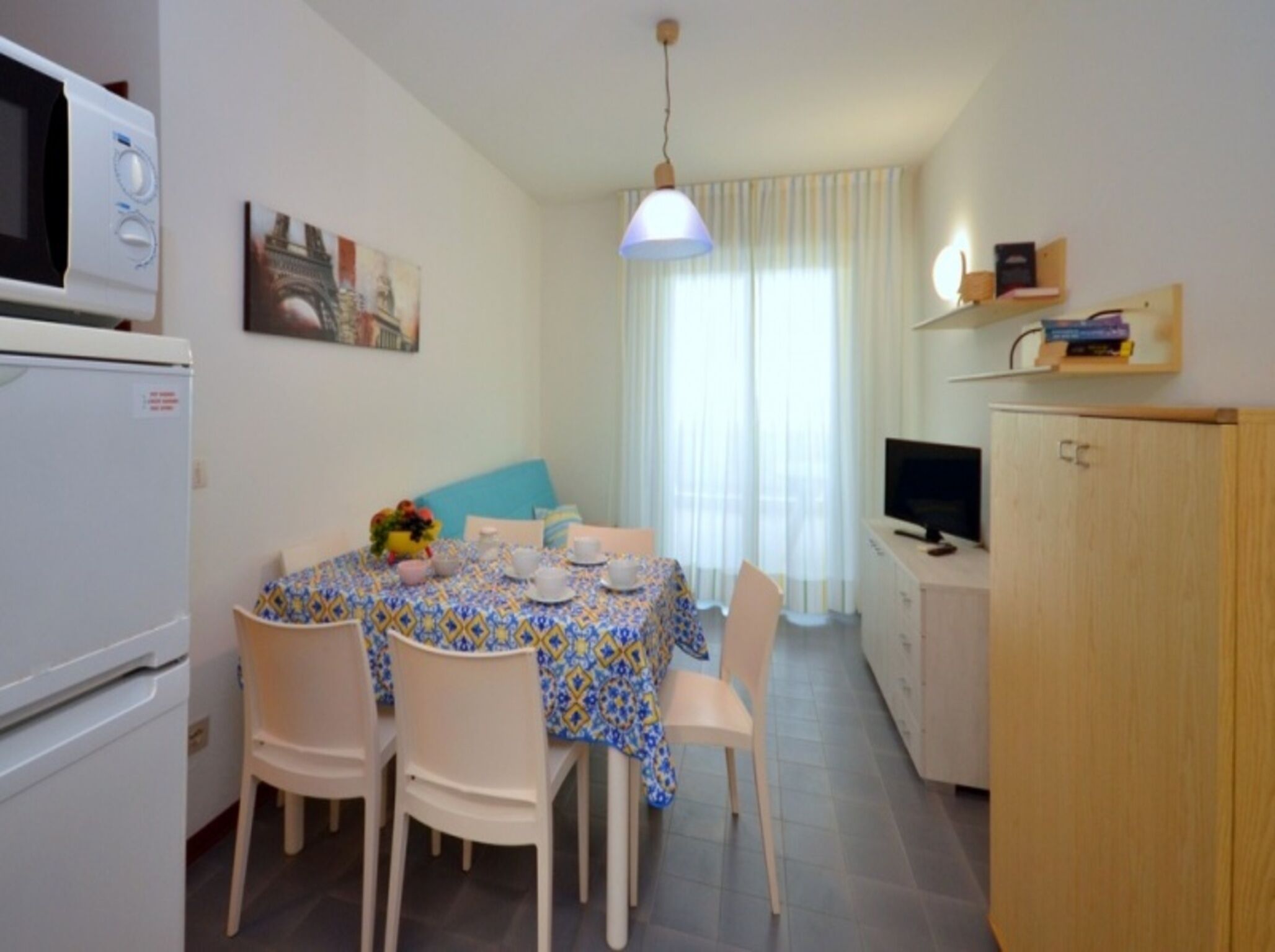 Un petit coin repas avec une table dressée pour quatre personnes, entourée de chaises blanches. Un four à micro-ondes est visible sur la gauche, et un téléviseur est posé sur un meuble en bois sur la droite. La pièce présente un aménagement simple et fonctionnel, avec un seul luminaire suspendu.