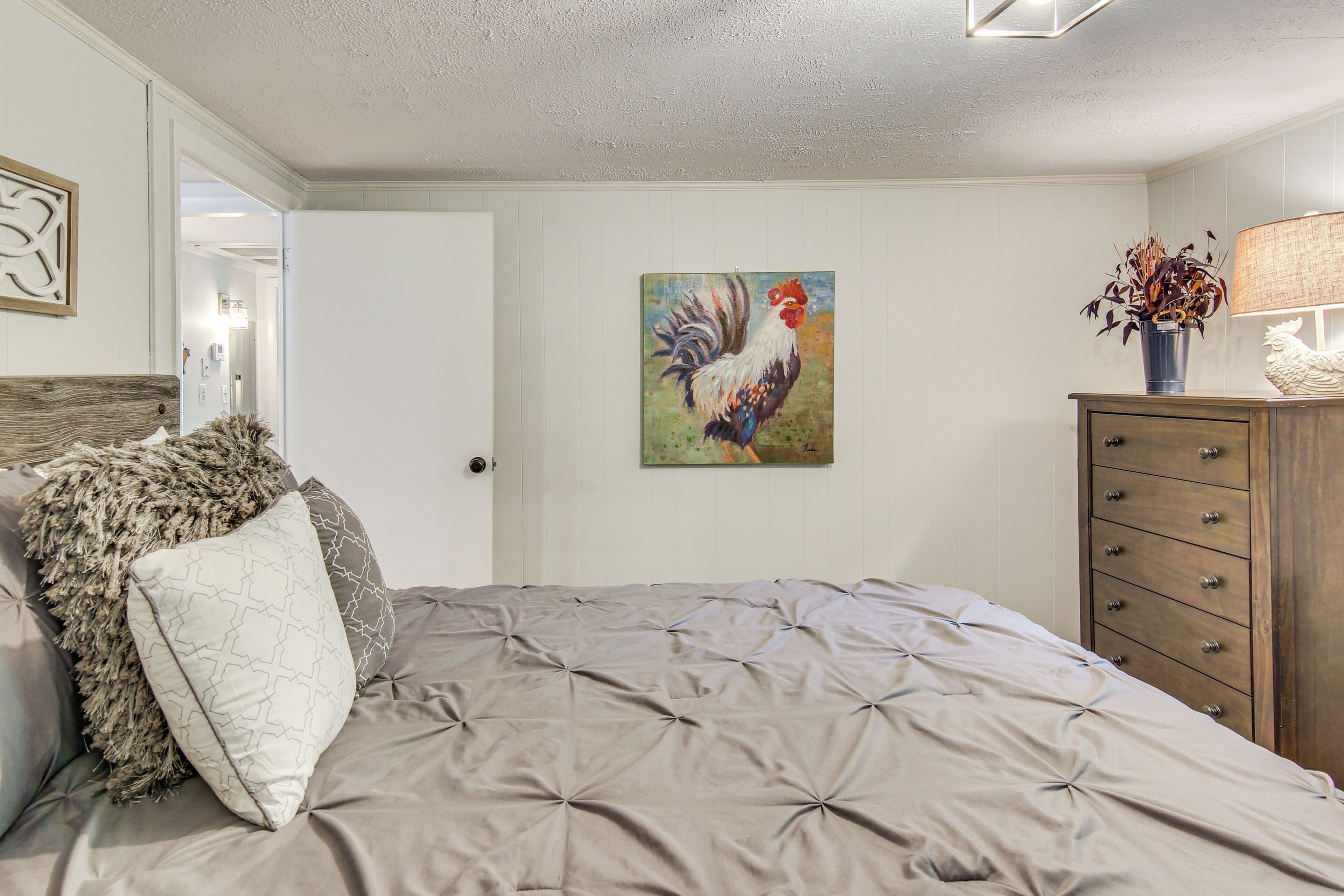 Un dormitorio con una cama, un aparador de madera, un cuadro de un gallo y una lámpara.
