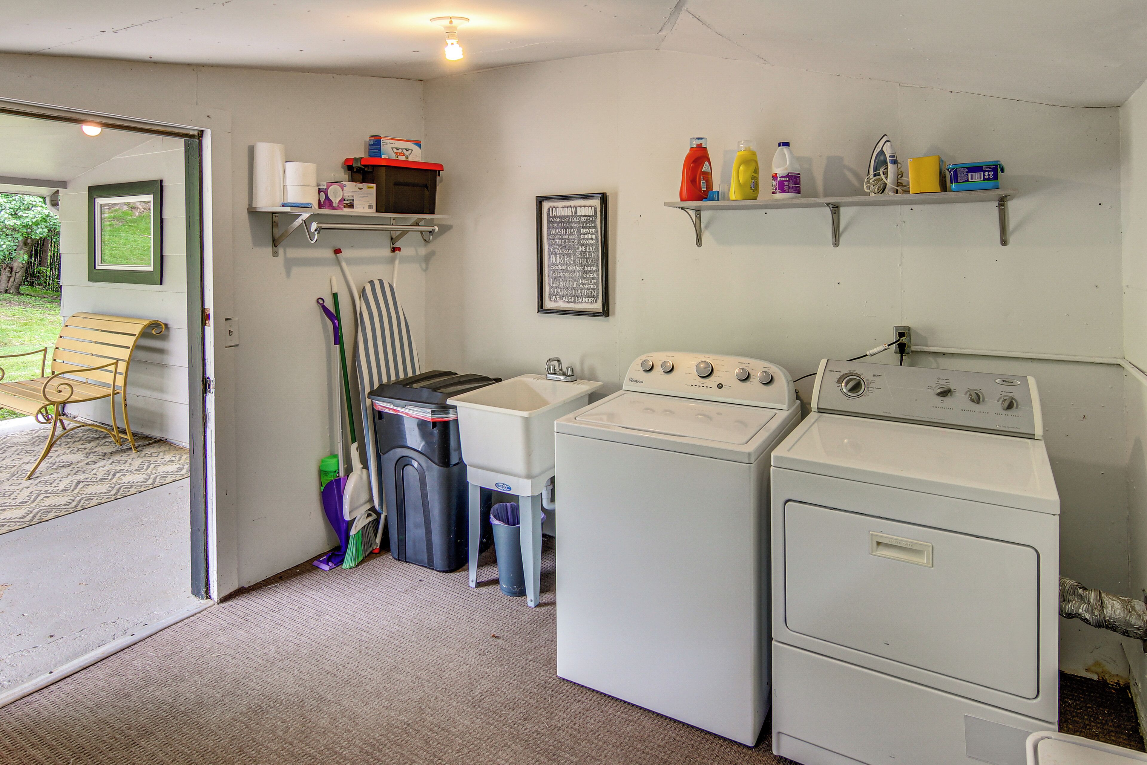 A laundry room with a washing machine, dryer, and ironing board. There are shelves with cleaning supplies and a door leading to an outdoor area.