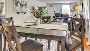 Un coin repas avec une table en bois, des chaises et un vase de fleurs. On aperçoit un canapé et une lampe au fond.