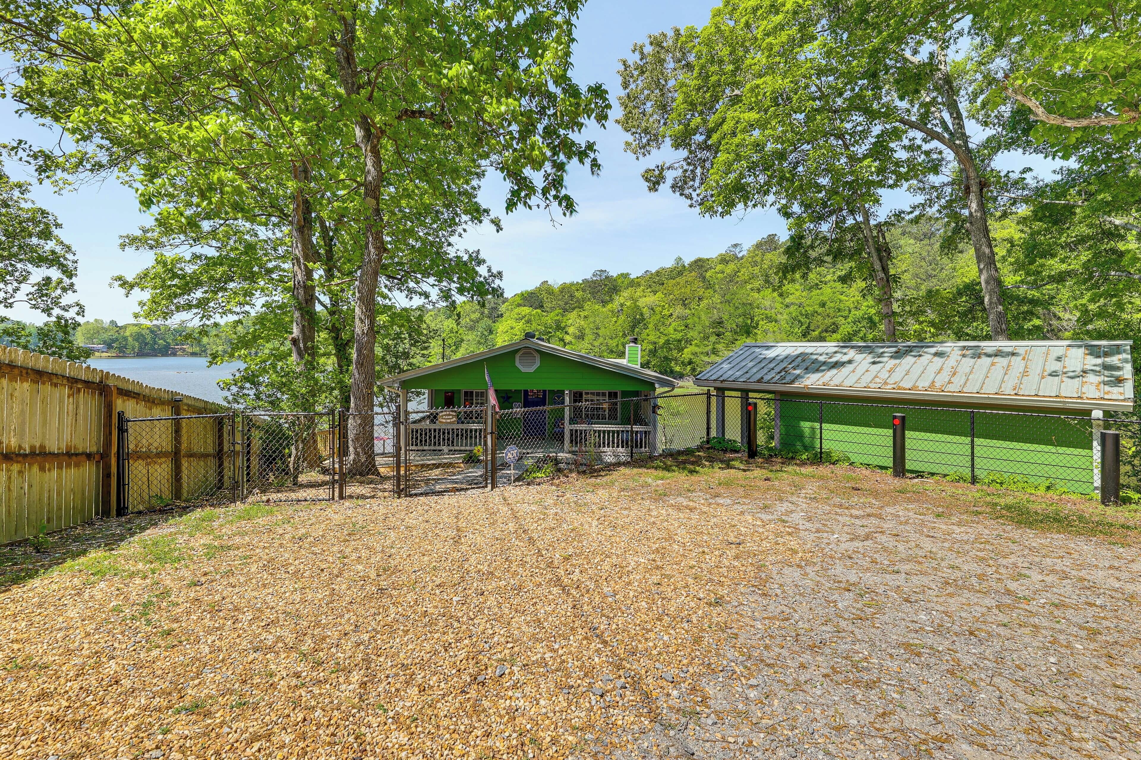 A gravel area with a fenced section, a green building with a porch, and a lake in the background.