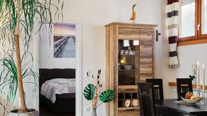 A room with a bed, a dining table with chairs, a wooden cabinet, a potted plant, and a wall-mounted shelf with decorative items. - Holiday Home 'Am Millibach' with Private Terrace, Private Garden and Wi-Fi (Wehingen)