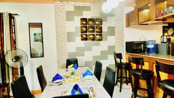 A dining area with a table set for four, black chairs, a fan, and a kitchen area with a microwave and refrigerator.