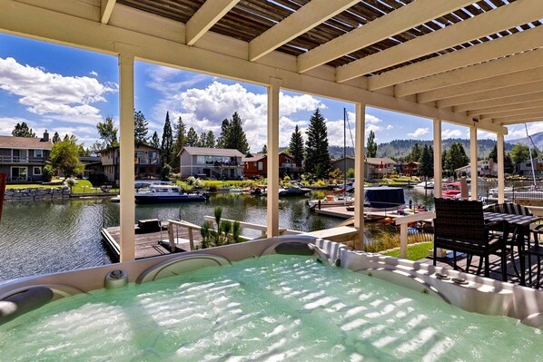 Jacuzzi con vista a un lago y barcos, rodeado por un porche cubierto con sillas y mesas.