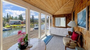 Une cabane dotée d’une grande fenêtre donnant sur un lac, avec un lit, une table ornée d’un vase de fleurs et une lampe.