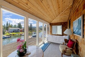 A cabin with a large window overlooking a lake, a bed, a table with a vase of flowers, and a lamp.