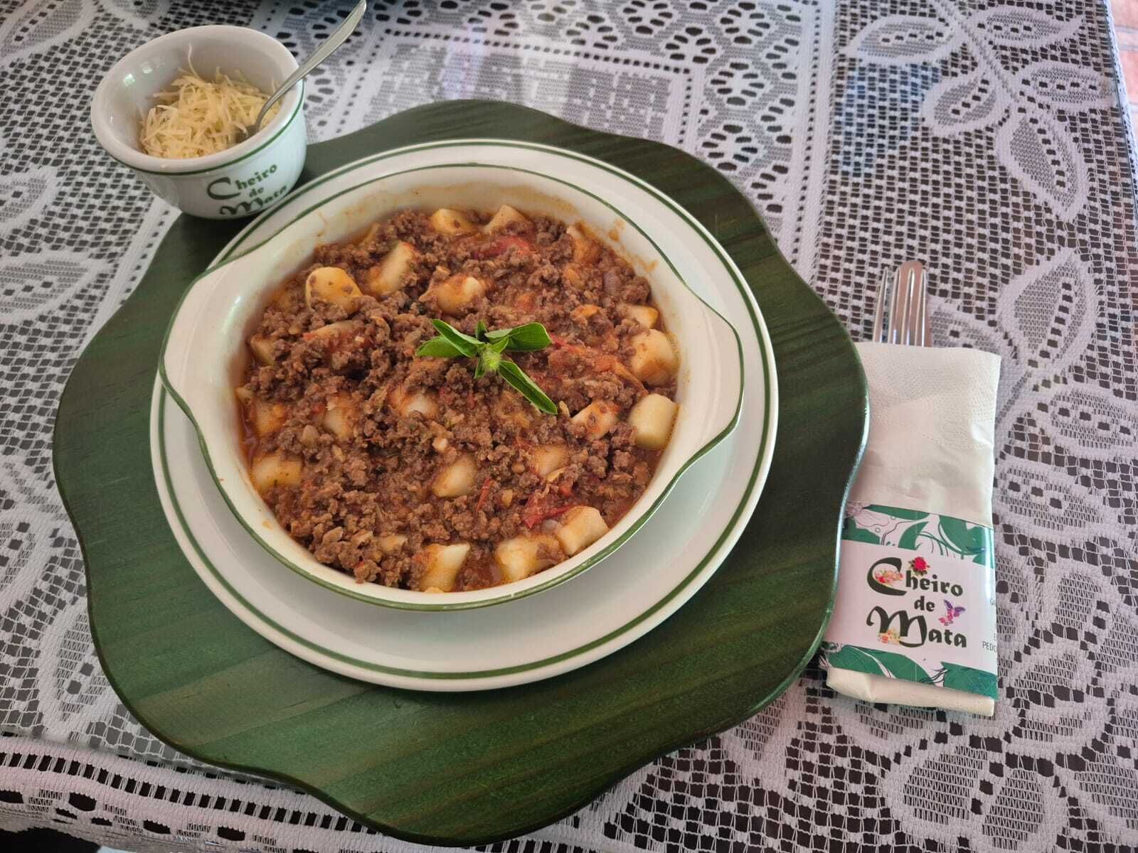 A bowl of pasta with a meat sauce, a small bowl of grated cheese, and a napkin with a logo on a table with a lace tablecloth.