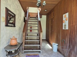 Una escalera de madera que lleva a un piso superior, una pequeña mesa con una maceta de barro, un cubo de basura y un aplique de pared.