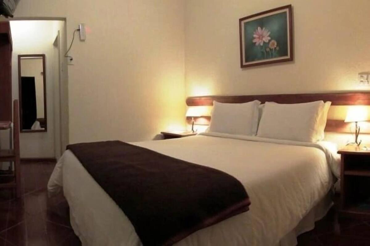 Ein Hotelzimmer mit Bett, Nachttischlampen, einem Nachttisch, einem kleinen Tisch und einem Bild an der Wand.