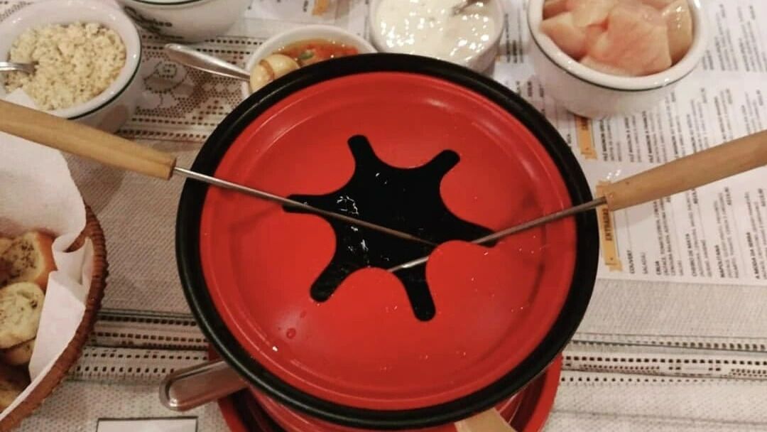 Un récipient rouge pour fondue, avec deux brochettes de nourriture, entouré de bols contenant diff érents aliments.