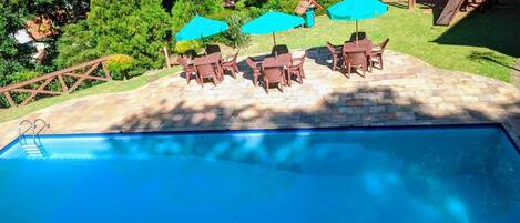 Una piscina con solárium de piedra, rodeada de árboles verdes y una estructura de juegos de madera.