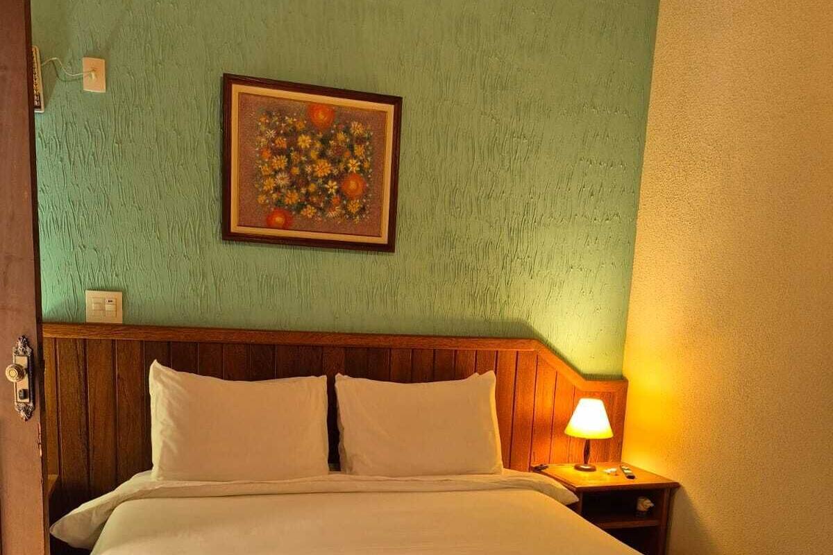 Ein Bett mit weißen Leinentüchern, ein Holz-Kopfteil, eine rote Tagesdecke, ein Nachttisch mit Lampe und ein gerahmtes Bild an der Wand.