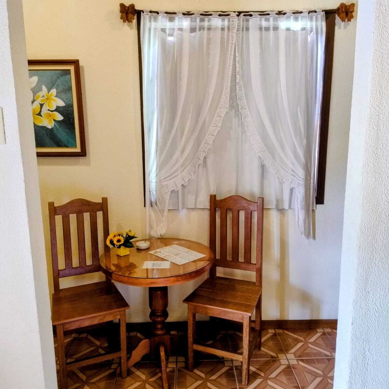 A small dining area with a round wooden table, two wooden chairs, a vase with sunflowers, and a framed picture on the wall.