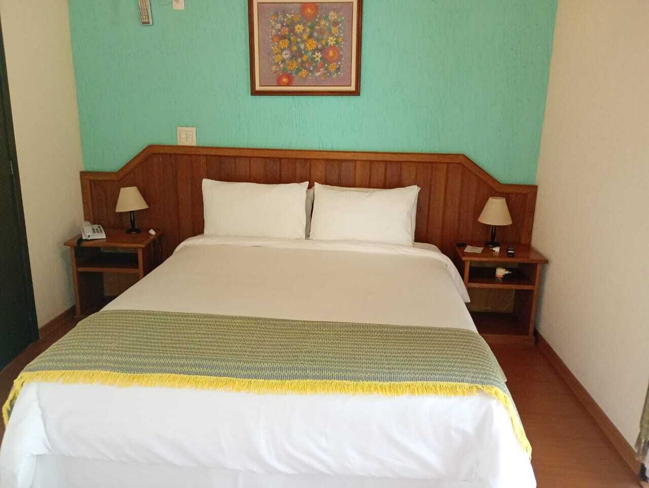 Una cama con cabecera de madera, dos mesitas de noche con lámparas, una obra de arte floral en la pared y una manta verde y amarilla a rayas.
