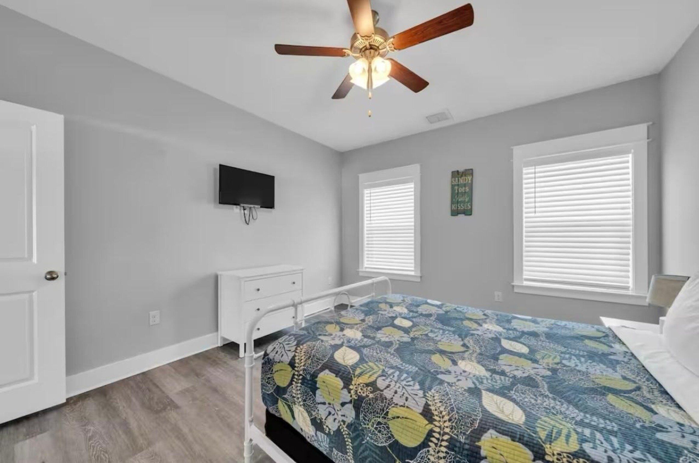 A bedroom with a bed, a ceiling fan, a TV, and two windows with blinds.