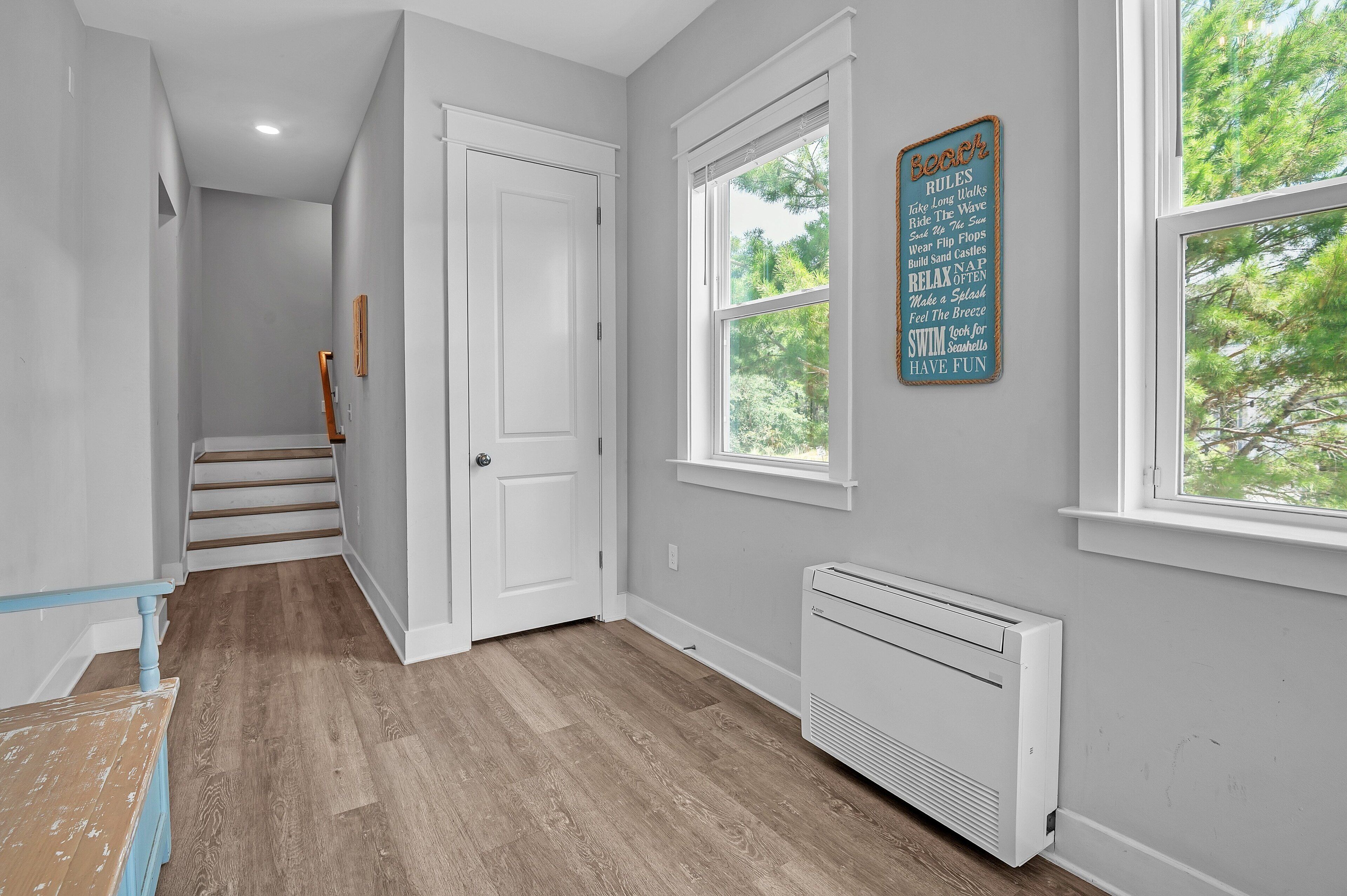 Un couloir avec un escalier, un radiateur blanc et une fenêtre donnant sur des arbres.