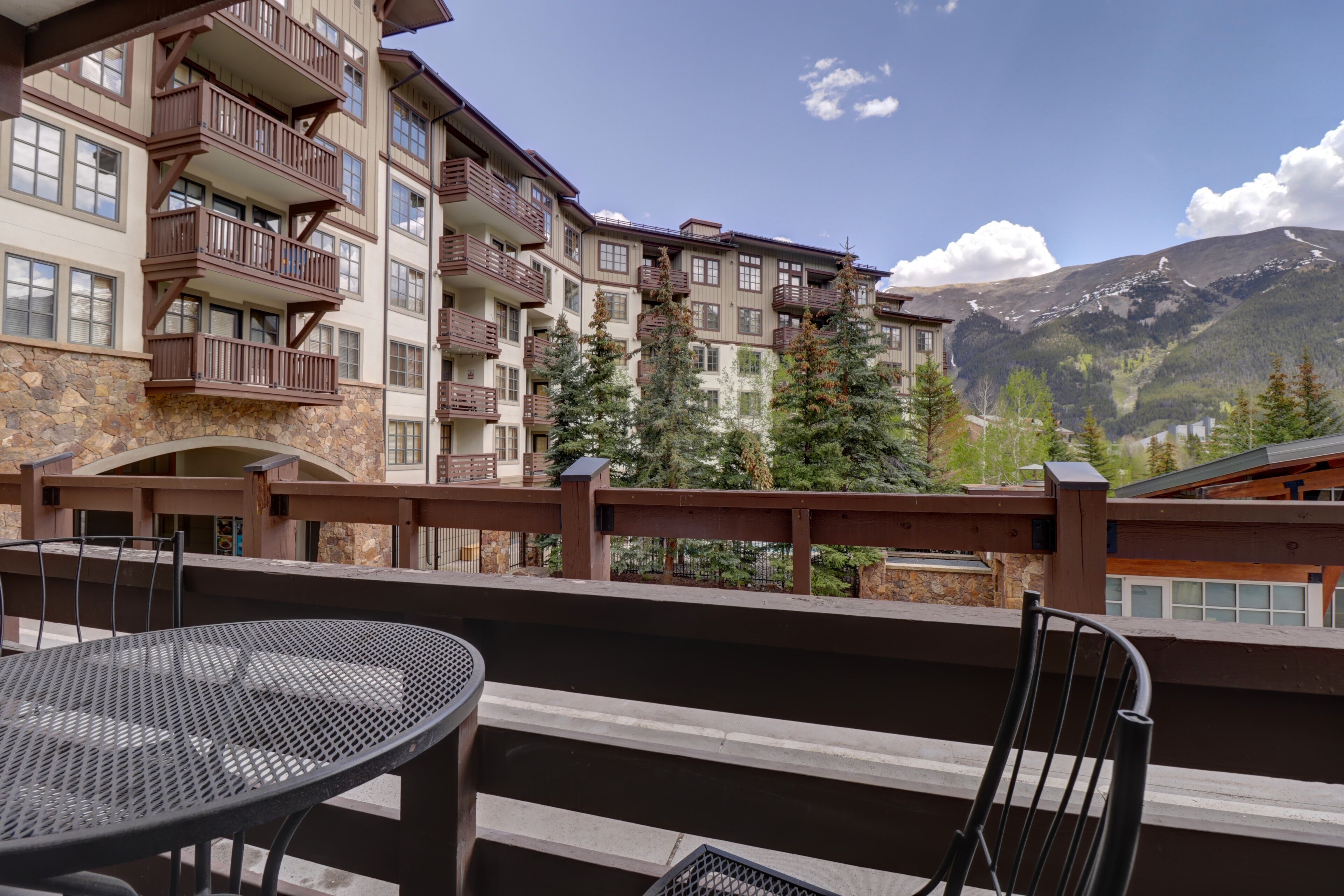 Un balcon d’hôtel avec une table et des chaises, donnant sur un paysage montagneux et un immeuble à plusieurs étages doté de balcons.