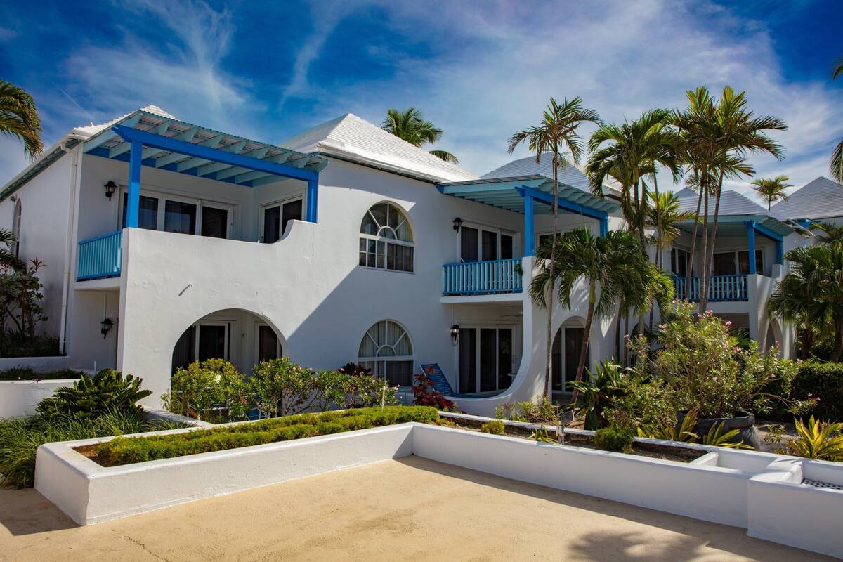 Un edificio blanco con balcones azules, ventanas arqueadas y un jardín ajardinado con palmeras.