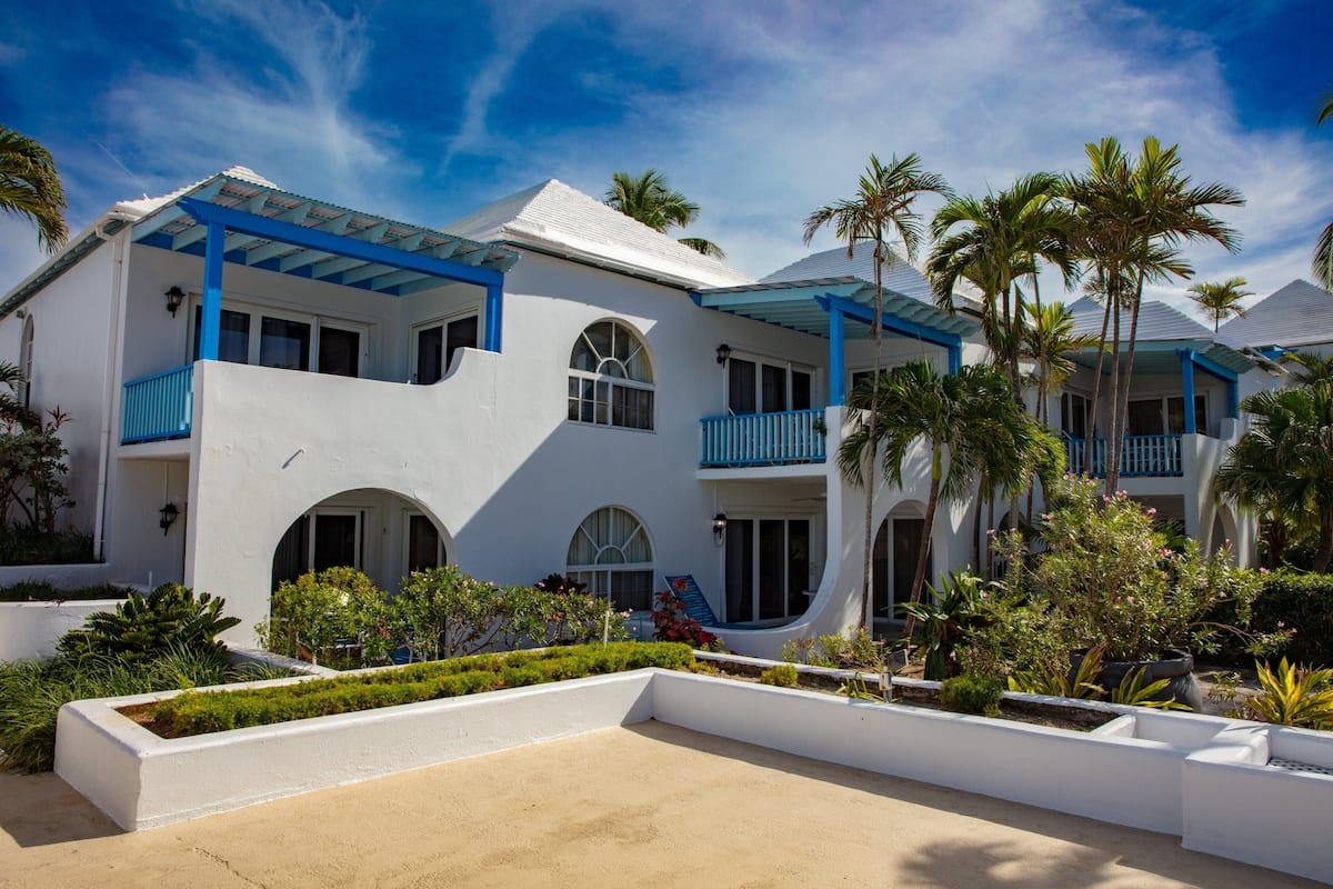 A white building with blue balconies, arched windows, and a landscaped garden with palm trees.