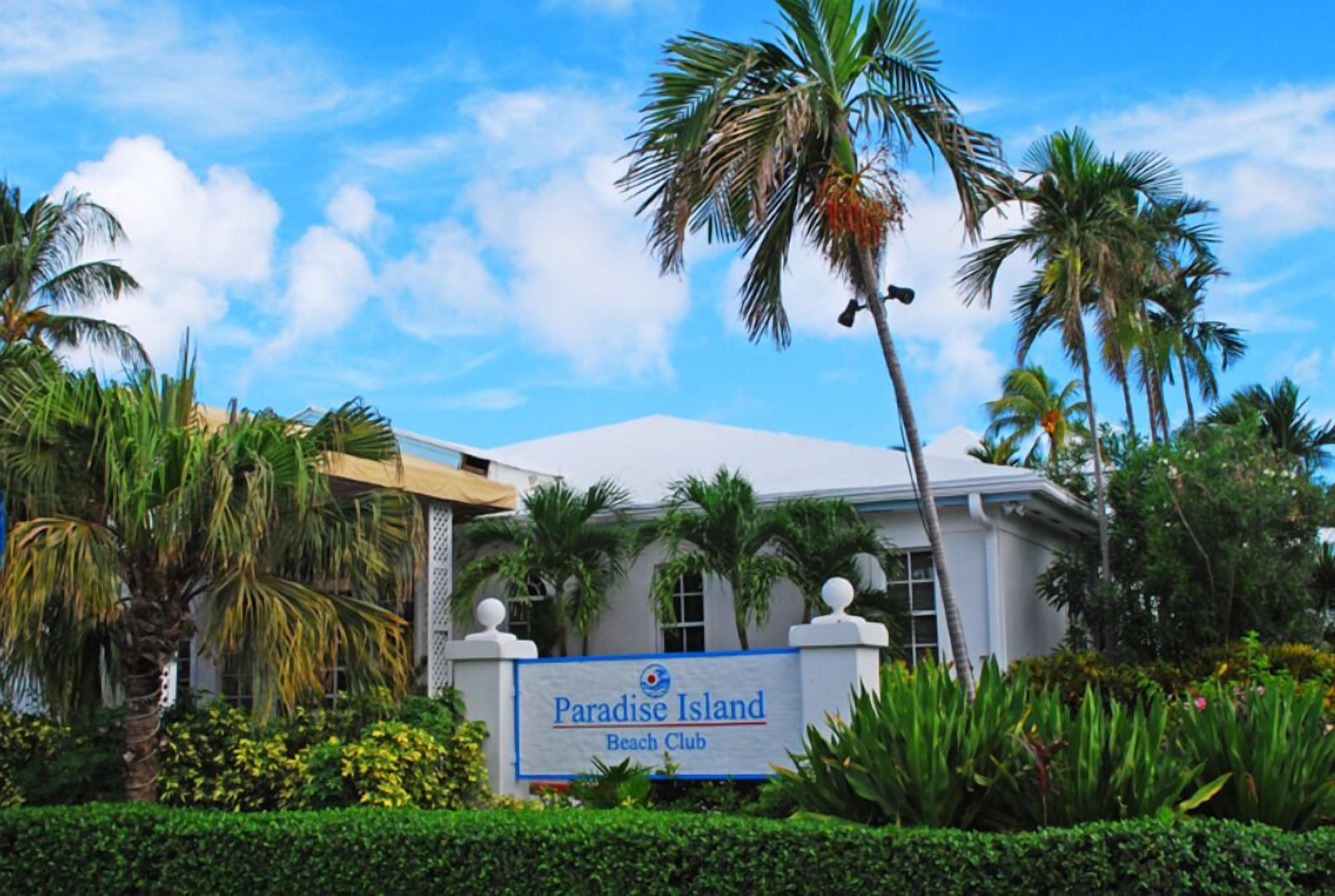 Ein Gebäude mit der Aufschrift "Paradise Island Beach Club", umgeben von Palmen und Grünpflanzen.