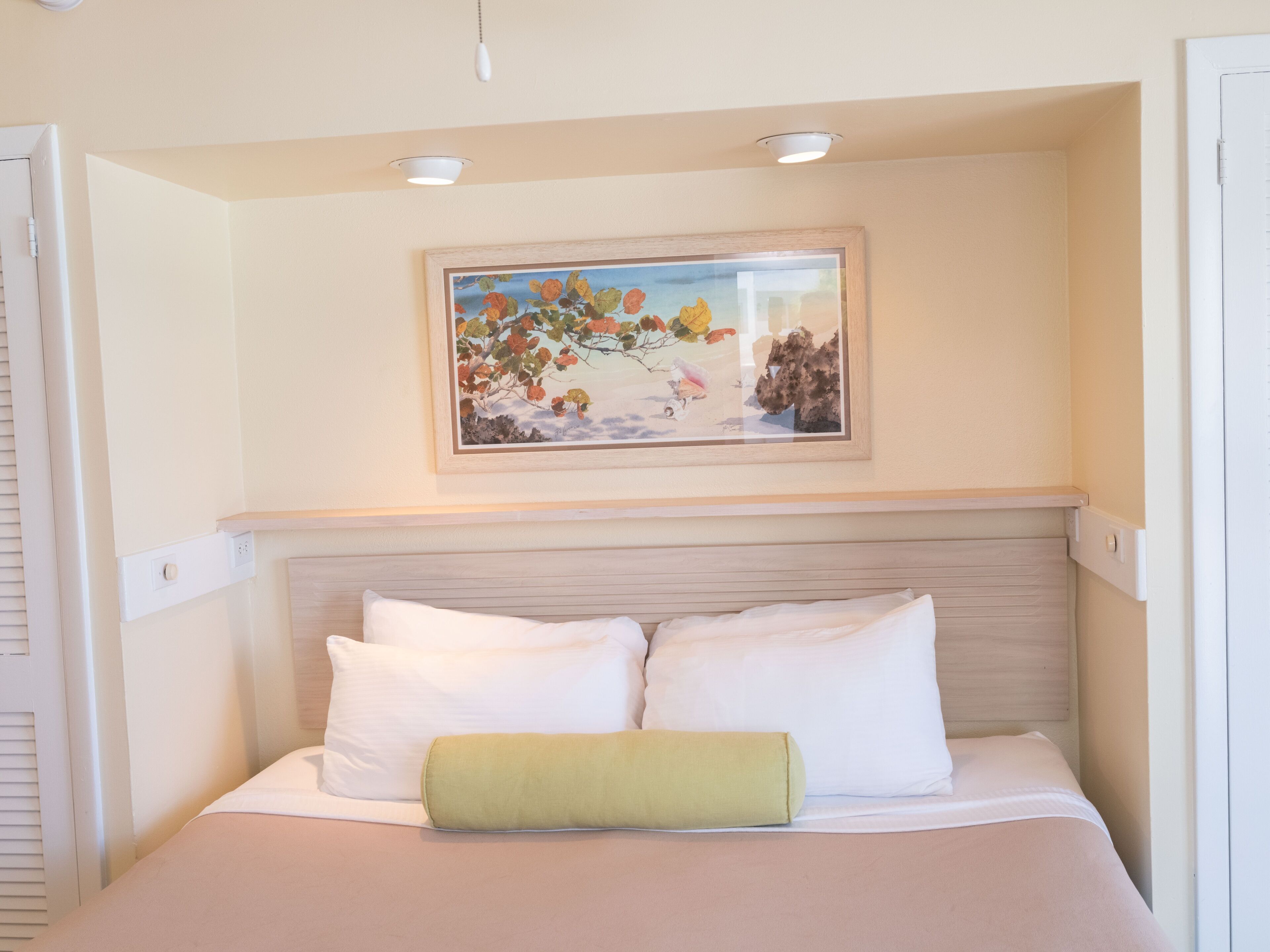 Un dormitorio con cama, almohadas blancas, una almohada verde, un cuadro enmarcado y luces empotradas en el techo.