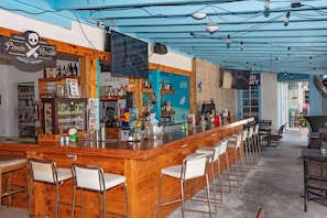 A bar area with wooden bar counter, multiple stools, and a variety of bottles and glasses. - Paradise Island Beach Club (Paradise Island)