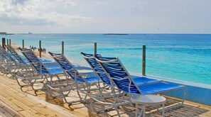 Fila de sillas reclinables azules sobre una terraza de madera con vista al mar azul y brillantes barcos a lo lejos.