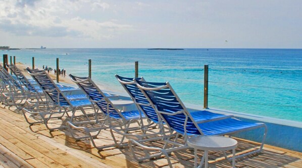 A row of blue lounge chairs with white metal frames on a wooden deck by the ocean.