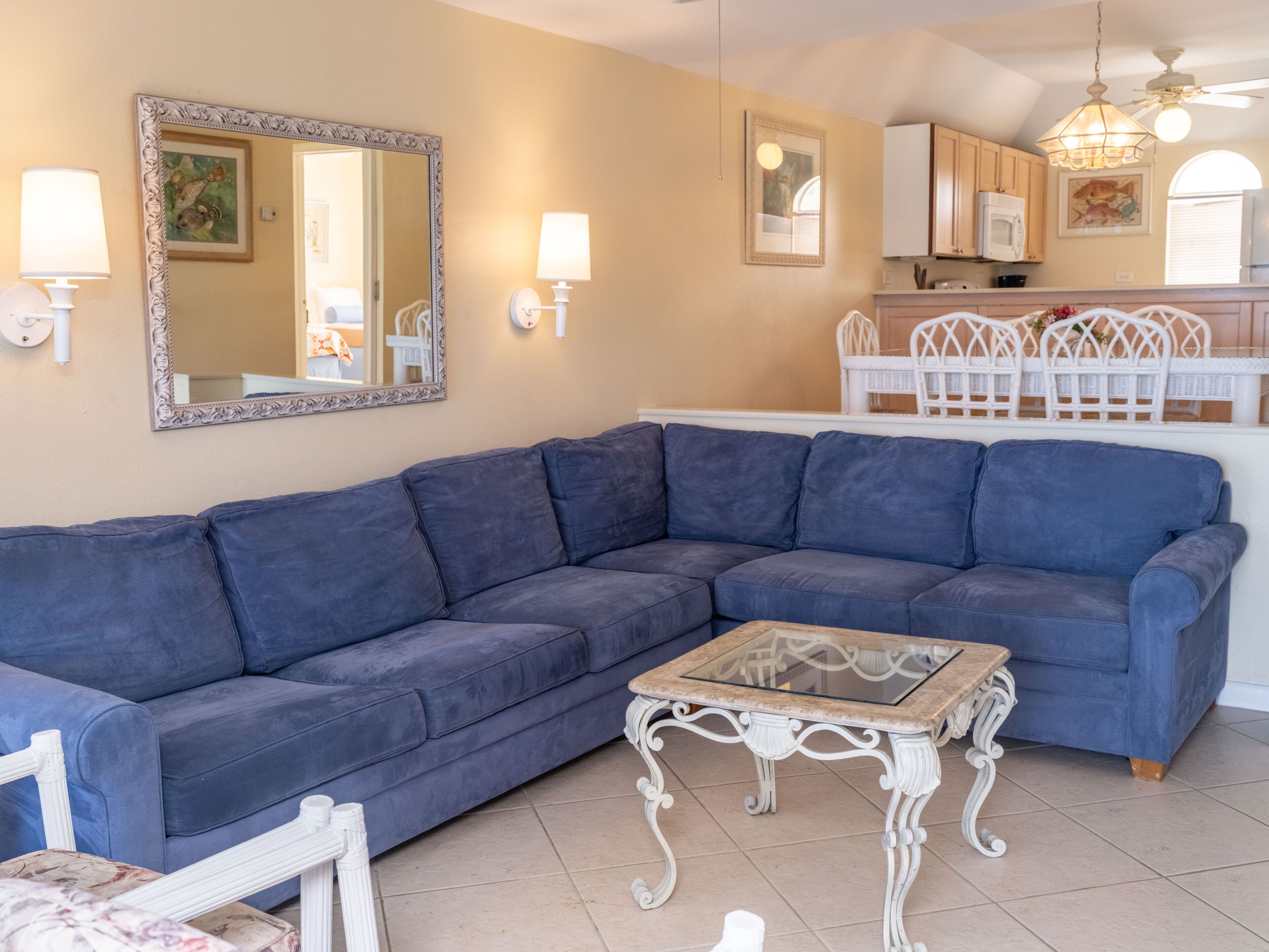 A living room with a blue sectional sofa, a glass-top coffee table, a white chair, and a kitchen area in the background.