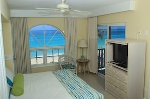 Una camera d'albergo con un letto, una televisione, una ventola a soffitto, una finestra vista oceano e un tavolino con una lampada.