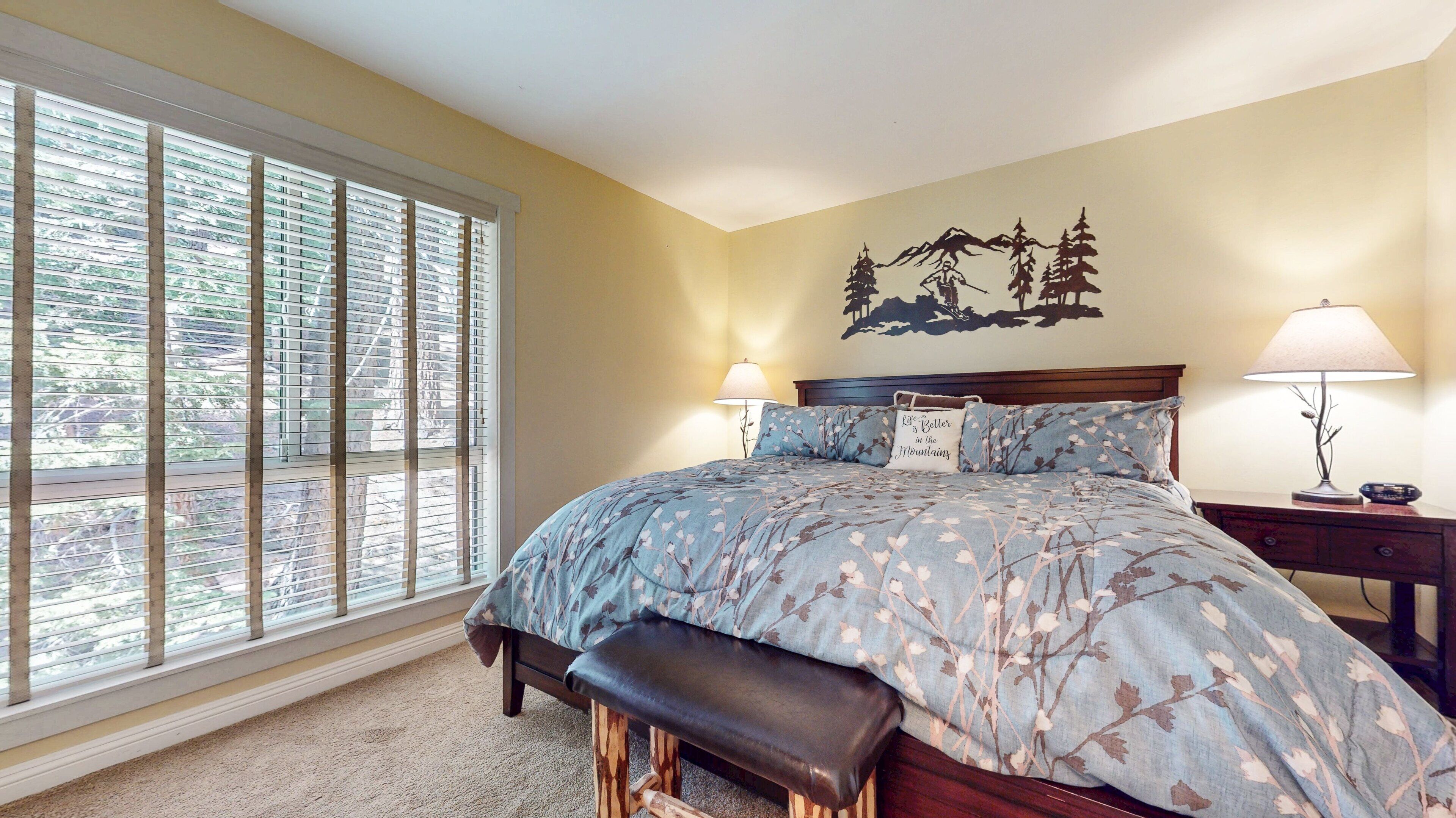 A bedroom with a large window, a bed with floral bedding, two bedside lamps, and a wall mural.