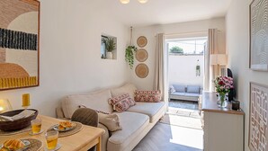 En hyggelig opholdsstue med sofa, spisebord med tallerkener og glas, en hængende lampe og udsigt gennem en dør til et udendørs område.