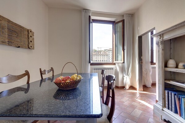 A dining area with a marble table, chairs, a fruit basket, a window with curtains, and a bookshelf with books.