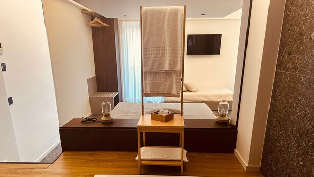 Ein modernes Hotelzimmer mit einer großen Badewanne, einem Nachttisch aus Holz mit Lampe, einem kleinen hölzernen Hocker, einem Bett mit Kopfteil und einem an der Wand montierten Fernseher.
