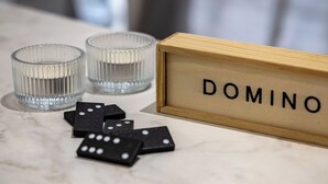 A marble table with a wooden nameplate labeled "DOMINO," two clear glass candle holders, and several black dominoes. - Nova Domo (Athens)