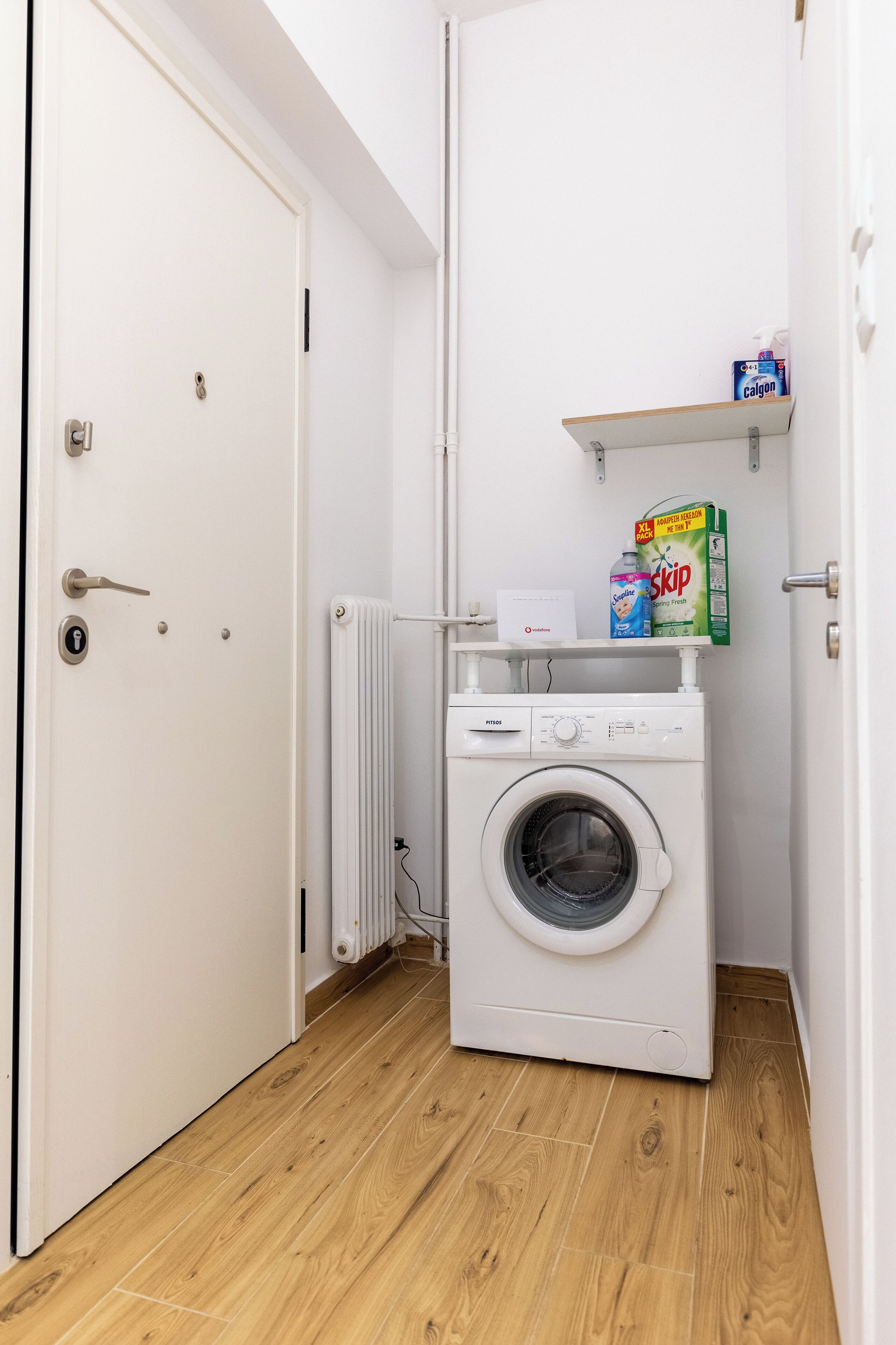 A compact laundry room with a white washing machine, a radiator, and shelves holding cleaning products.