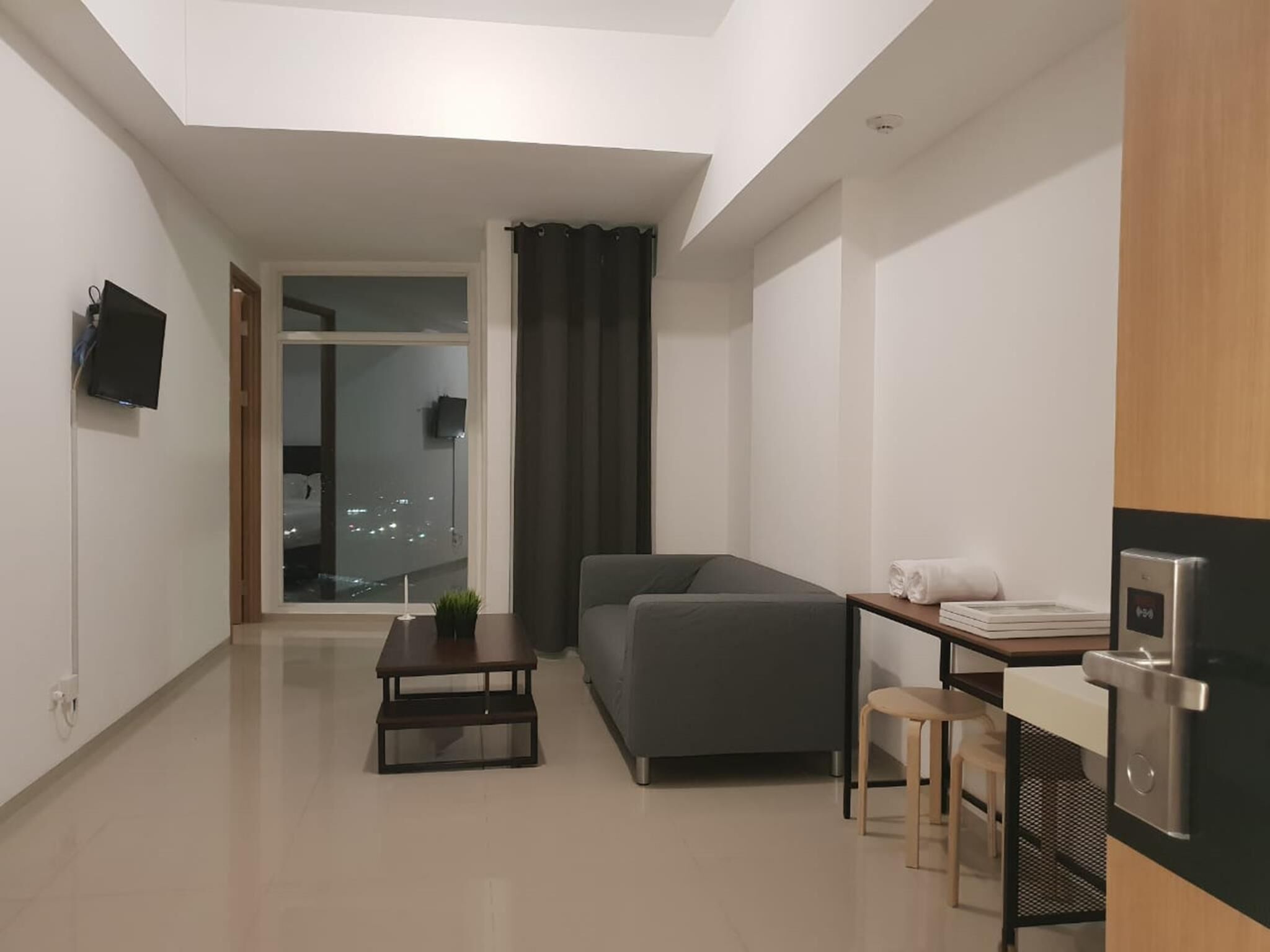 A modern, minimalistic living room with a grey sofa, a small coffee table, a TV mounted on the wall, and a view of the city through a large window.