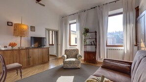 A living room with a sofa, armchair, wooden cabinet, television, and a view of the city through large windows. - Green Dragon - Bright apartment with Arno view (Firenze)