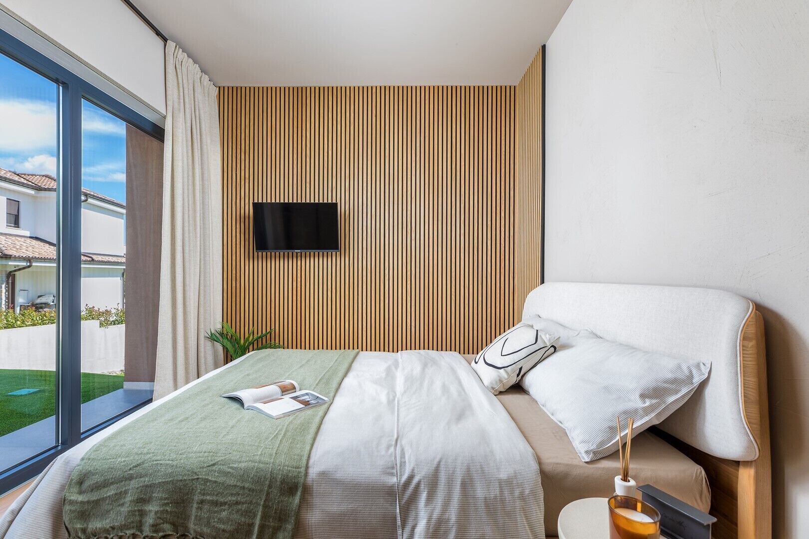 Een moderne hotelkamer met een groot raam, een bed met een groene deken, een nachtkastje met een lamp en een aan de muur bevestigde televisie.