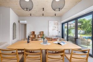 Ein geräumiger Essbereich mit einem großen Holztisch und Korbstühlen, einem modernen Deckenbau und Blick auf einen Garten und Pool durch große Fenster.