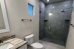 A bathroom with a marble countertop, a toilet, a glass-enclosed shower, and a small window.