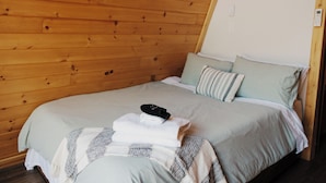 A neatly made bed with a folded towel and a black object on top, set against a wooden wall.