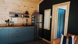 A compact kitchen with a stainless steel refrigerator, wooden shelves with glassware, and a brown leather couch.
