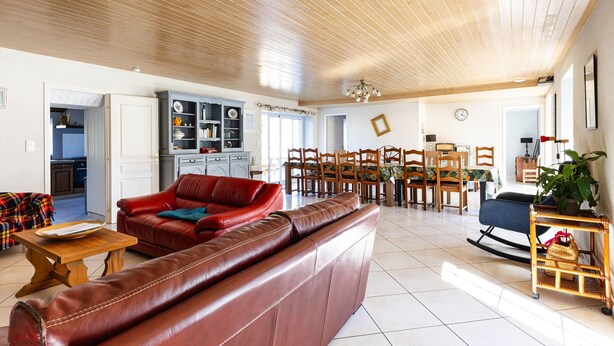 A spacious living room with a wooden ceiling, a red leather sofa, a dining area with wooden chairs, and a kitchen visible through an open door. - Gite O Pré'Vert, Gîte Ô Pré'Vert. Group gite, tourist rental in Le Poiré Sur Vie in the Vendée. G... (Le Poiré-sur-Vie)
