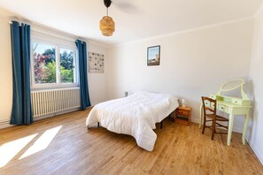 Un dormitorio con cama, escritorio, silla, radiador, ventana con cortinas y un cuadro en la pared.