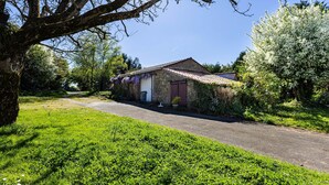 A stone building with a tiled roof, a paved driveway, and lush greenery. - Gite O Pré'Vert, Gîte Ô Pré'Vert. Group gite, tourist rental in Le Poiré Sur Vie in the Vendée. G... (Le Poiré-sur-Vie)